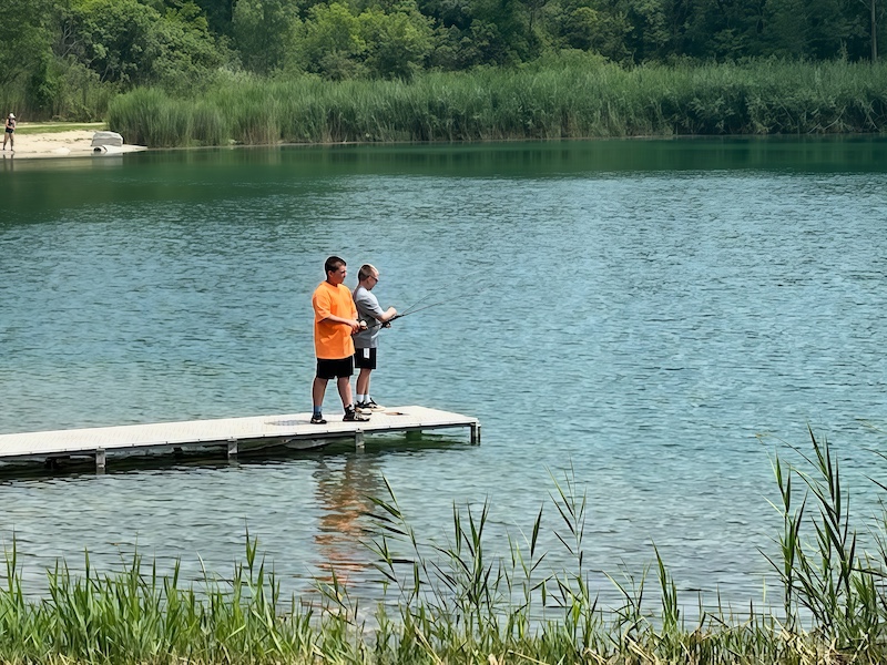 Two children stand on a dock on a pond and try to catch fish with rods and reels. In the background is a shoreline filled with tall grasses against a forest.