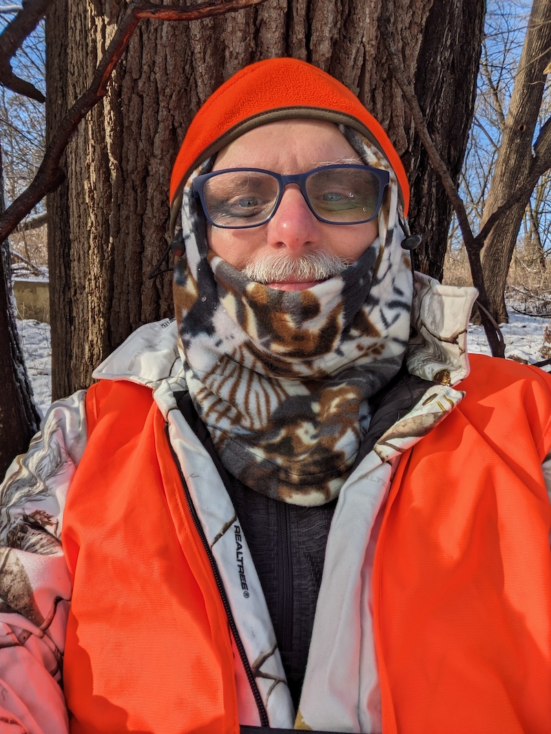 A portrait of a hunter sitting on the ground leaning against a tree trunk wearing blaze orange and snow camouflage. In the background is a snowy wooded landscape.
