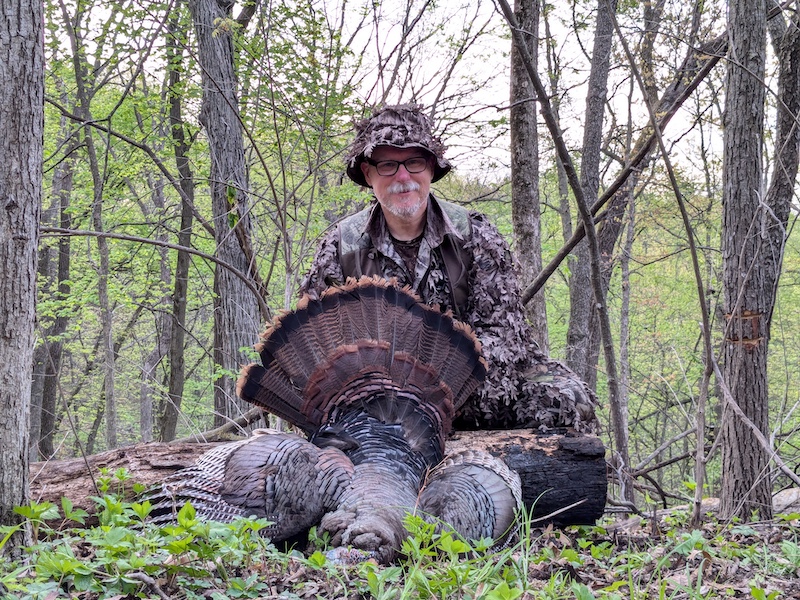 In an early spring woodland, a hunter wearing camouflage is kneeling on the ground and posing with a successfully harvested wild turkey.