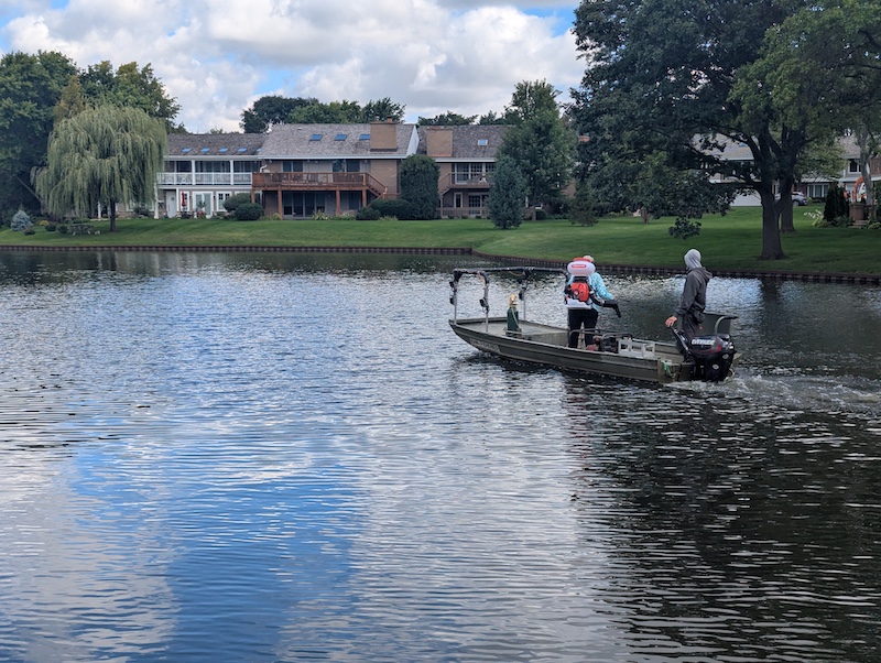 Two people stand in a boat as it moves across a waterway in a subdivision. On the boat, one man runs the motor in the back, and the other man applies herbicide with a backpack blower.