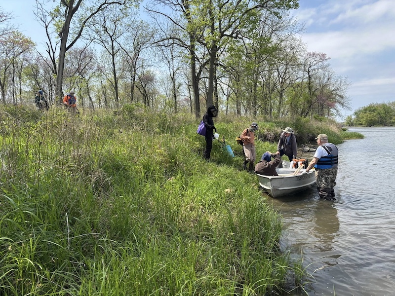 A group of people walking down towards a small boat on the shoreline of a river. The people grab supplies from the boat to aid in habitat conservation efforts. One person wearing waders and a life vest holds the boat to the shoreline while wading in the river.