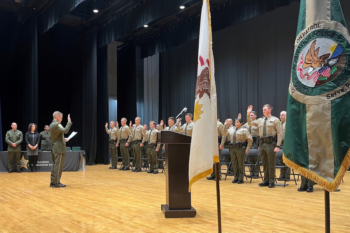 A group of conservation police officers standing in a group on a stage with black curtains in the background. The group is all wearing officially uniforms including olive green pants, tan shirt and an olive green tie. The officers are all holding up their right palms as if they are being sworn in. A senior officer in all olive green stands in front of the group reading off a sheet of paper and has his right palm facing out toward the officers. Three officials stand to the right and off to the side of the stage and observe the proceedings.