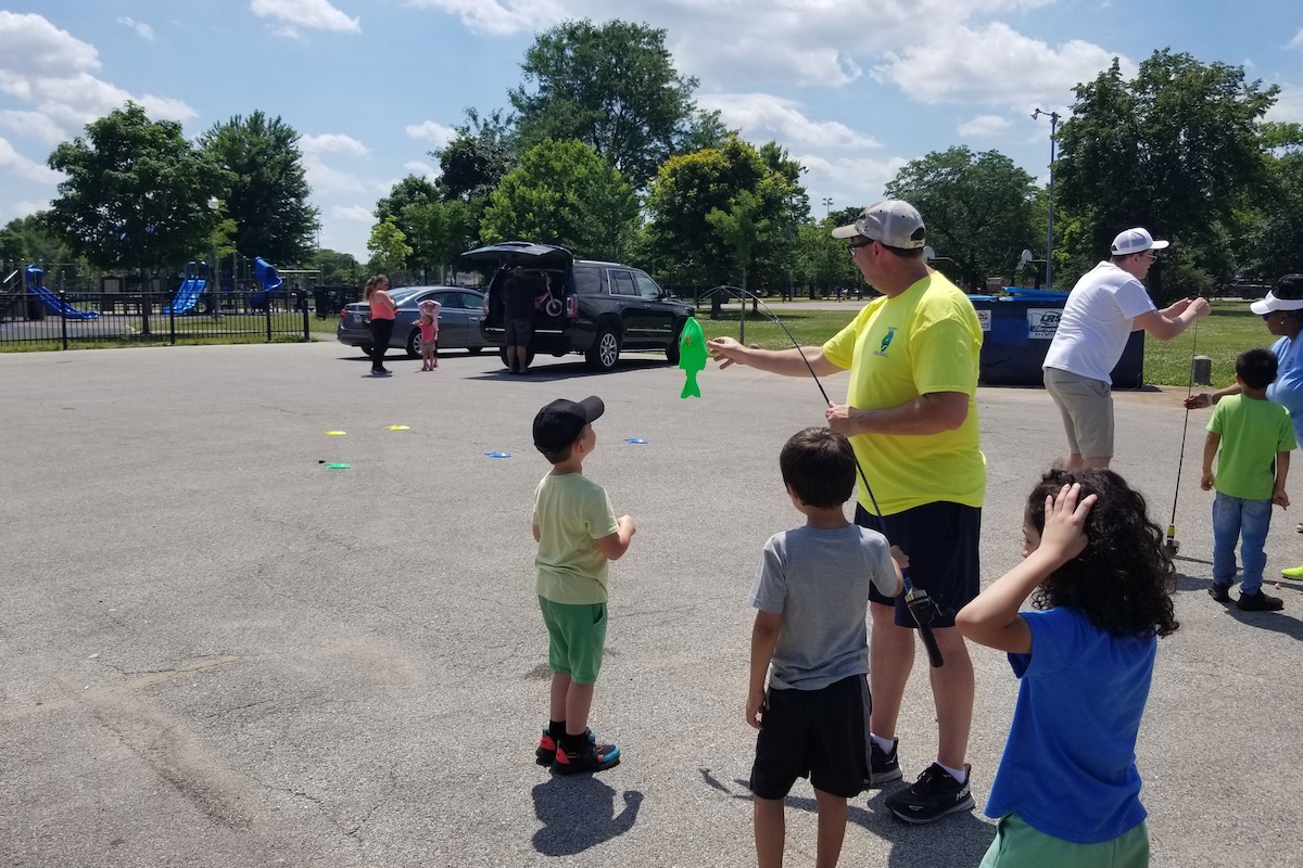 Two adults demonstrate to young children how to use a fishing rod and reel with plastic fish on the ends of the line.