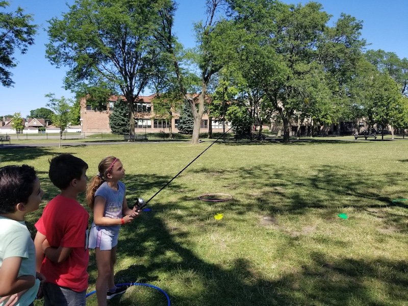 On a sun dappled lawn, one child holds a fishing rod and reel while two other children wait for their turn. In the background is a brick two story building surrounded by large trees.