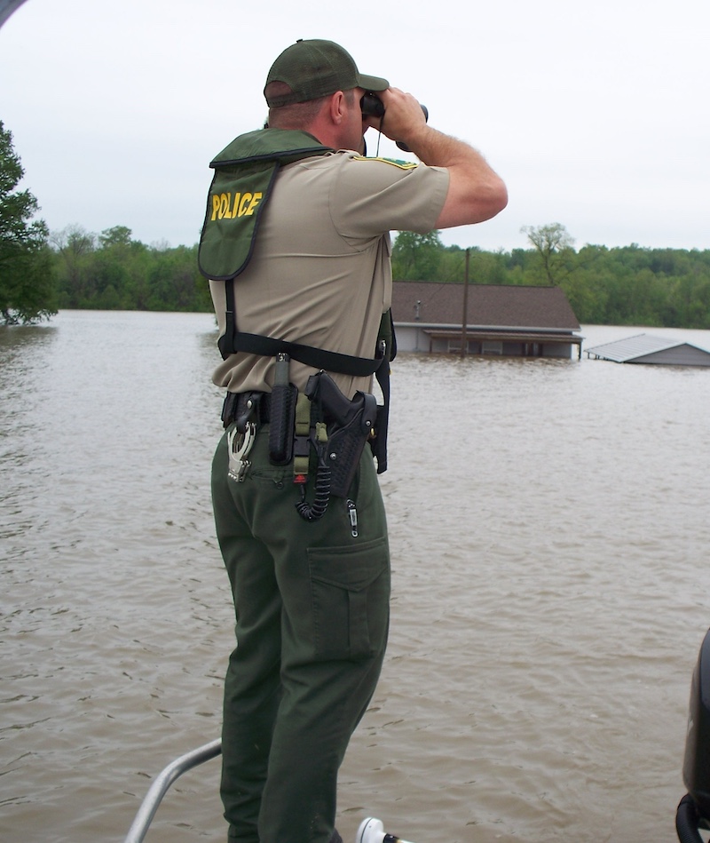 A conservation police officer stands on a boat and looks through binoculars at flood damage. In the background a house nearly submerged in water with just its roof above water.