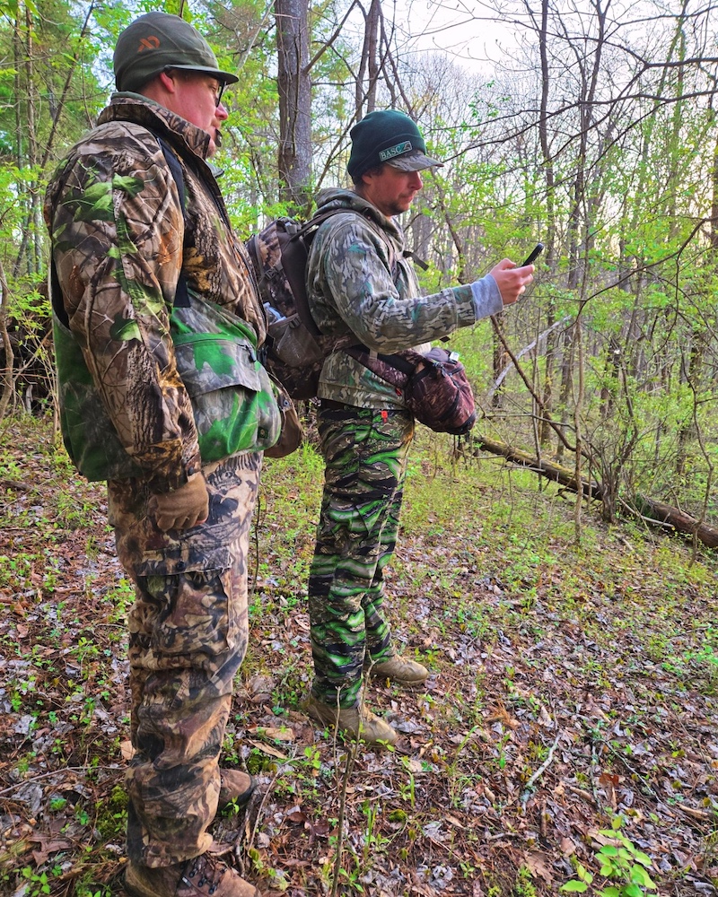 Two hunters wearing camouflage gear stand in a a woodland. One hunter holds up his phone and they both look at its screen. In the background sunlight is peeking through trees that are just beginning to leaf out in an early spring morning.