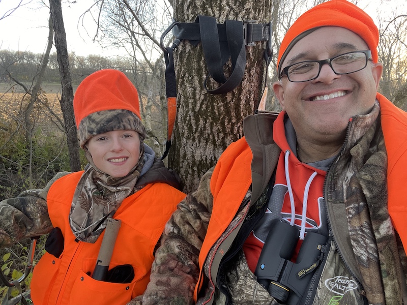 An adult and youth hunter wearing blaze orange sit in a tree stand. In the background is an agricultural field with a fencerow of trees.