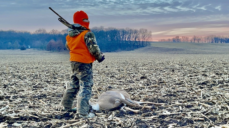 In an agricultural field, a hunter stands next to a successfully harvested white-tailed deer. In the background is the sunrise over a wooded hilltop.