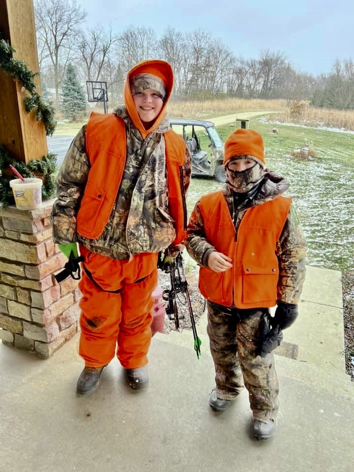 Two young hunters wearing blaze orange gear stand together on a front porch. In the background is an ATV parked on a grassy lawn next to a gravel driveway.