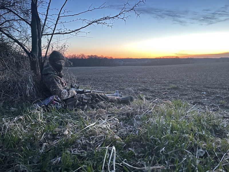 A hunter in camouflage gear sits at the base of a tree on the edge of an agricultural field at daybreak. The hunter rests his shotgun on his lap.