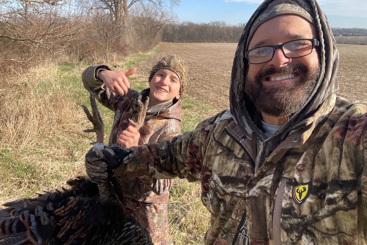 An adult and youth hunter stand on the edge of an agricultural field. They both hold up a successfully harvested wild turkey. Both are wearing camouflage gear.