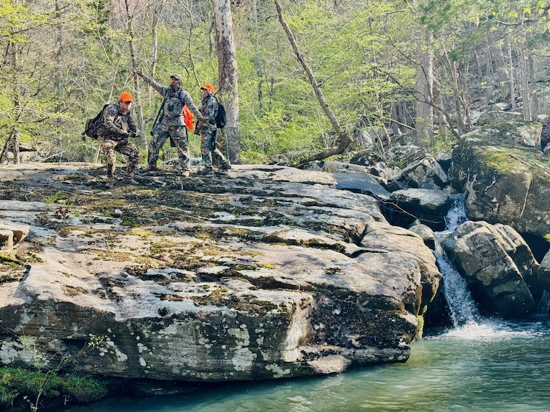 Three hunters stand on a boulder next to a stream and pose for the camera. All three are wearing camouflage gear. In the background is a spring woodland.