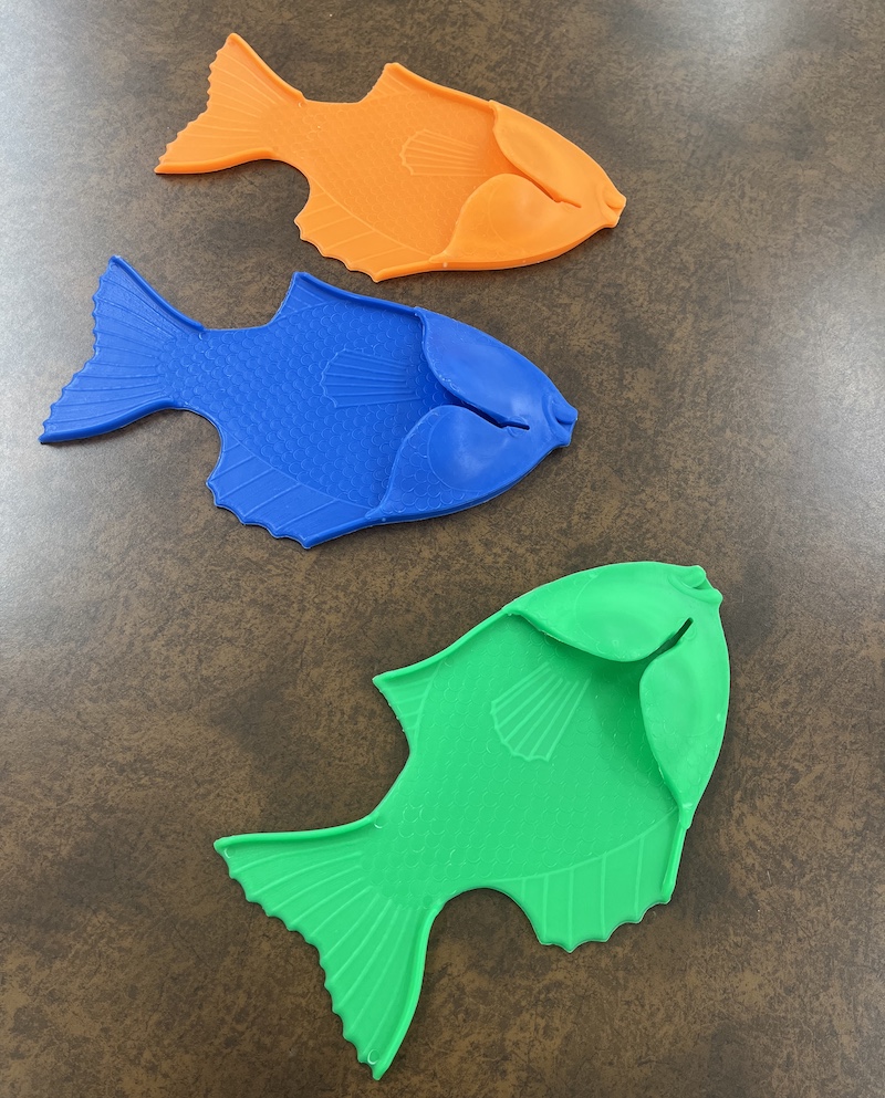 Three plastic fish rest on a counter top. One fish is orange, another is blue and the last is green.