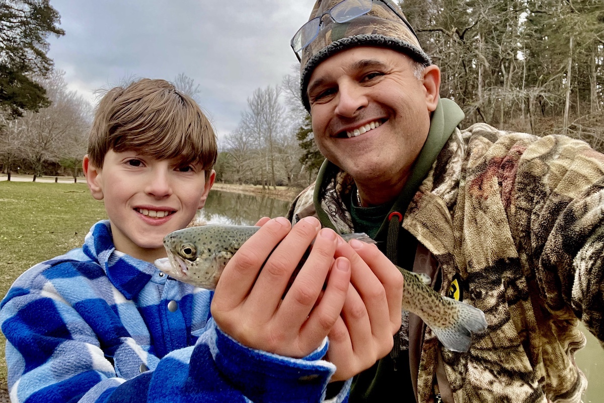 A father and son take a selfie while the son holds up a trout in both hands. In the background is a pond under a overcast sky.