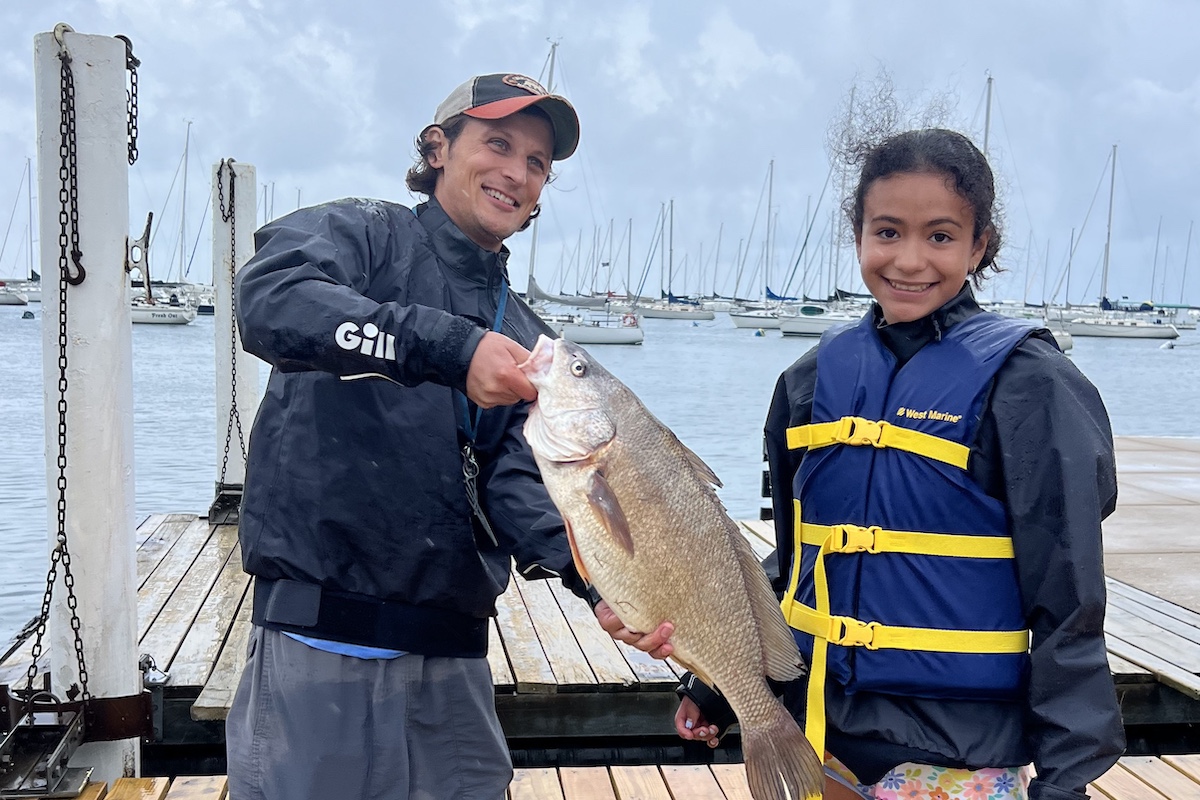 A young girl smiles excitedly while standing next to a man who is holding a large gray fish with both hands. Both are wearing wet weather jackets. The girl is wearing a life-vest. In the background is a wooden dock over lake with many sail boats anchored nearby.