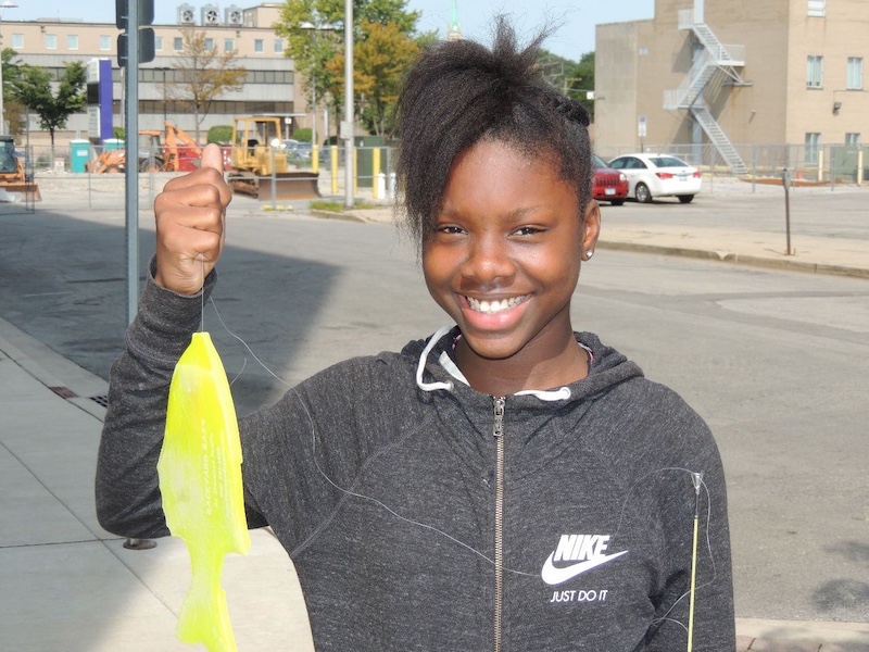 A child holds up a yellow plastic fish successfully caught on a fishing line. In the background is an urban parking lot and tan brick buildings.