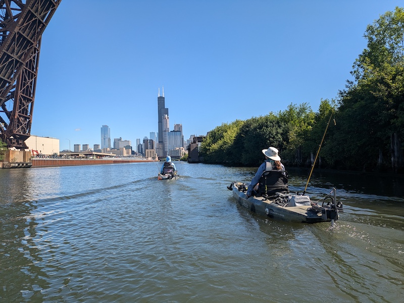 Two people float on the Chicago River on kayaks. Each kayak has a fishing rod and reel secured to its side. In the foreground is the Chicago skyline. A bright blue sky is overhead.