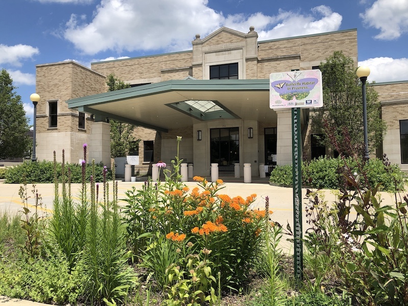 A flowerbed of various native prairie plants blooms in front of a tan building. There is a sign on a green pole in the flowerbed saying, "Butterfly Habitat in Progress." Above is a bright partly cloudy sky.