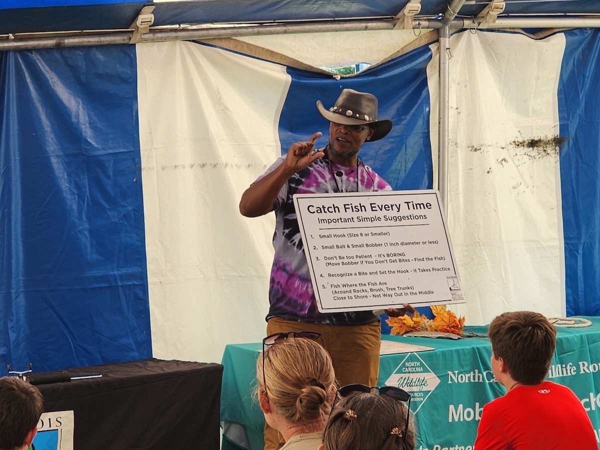 Eliha Perez at a fishing event teaches students tips for catching fish. The sign he is holding says: Catch Fish Every Time.