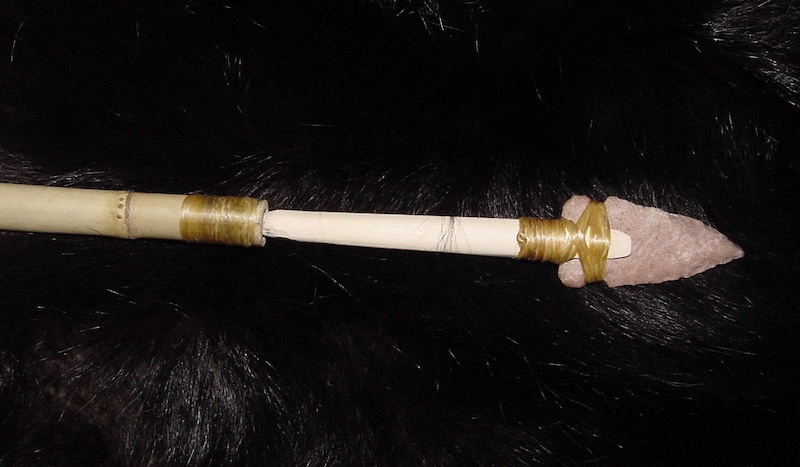 A stone arrowhead point is attached firmly to the shaft of an atl-atl spear. The spear is resting on black fur.