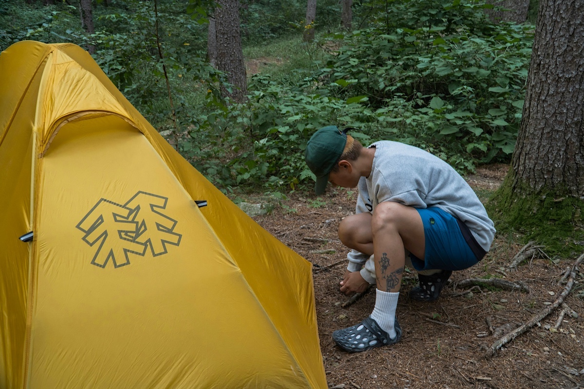 A young man secures his yellow tent with a rope tied to a stake. In the background is the green understory of a woodland.