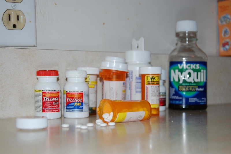 A line of assorted prescription pills and medicine bottles line up on the back of a counter-top. In front of the line is a pill bottle on its side with its cap off and capsules spilled out on to the counter.