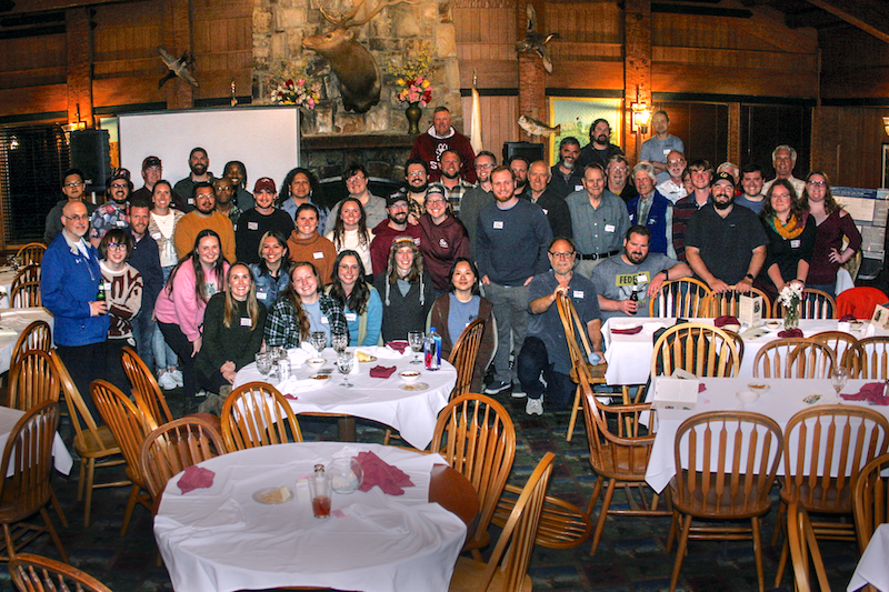 A group of people stand together in front of a banquet hall with tables covered in white linens surrounded with wooden chairs. In the background is a large fireplace with a projector screen.