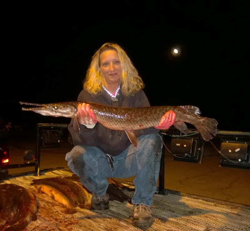At night, an angler squats in a trailer as she holds up proudly her large fish successfully harvested for the table. 