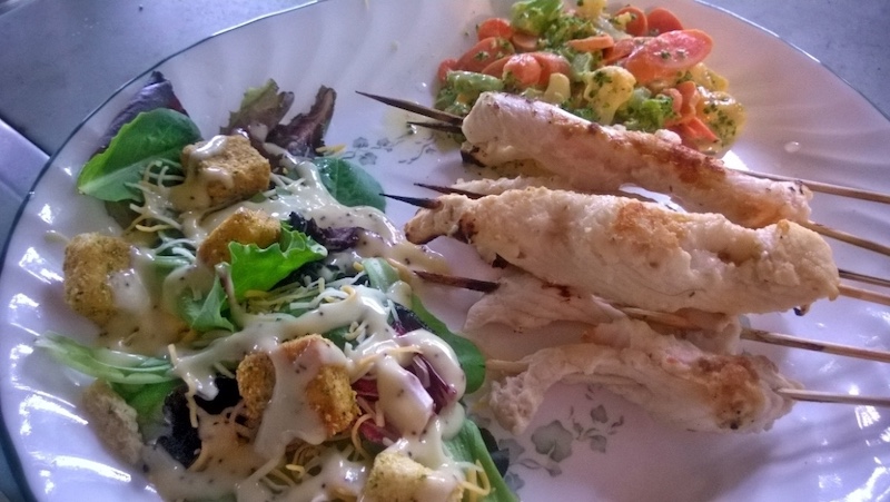 A plated meal of fish grilled on skewers along with a green leafy salad and a side of mixed vegetables.