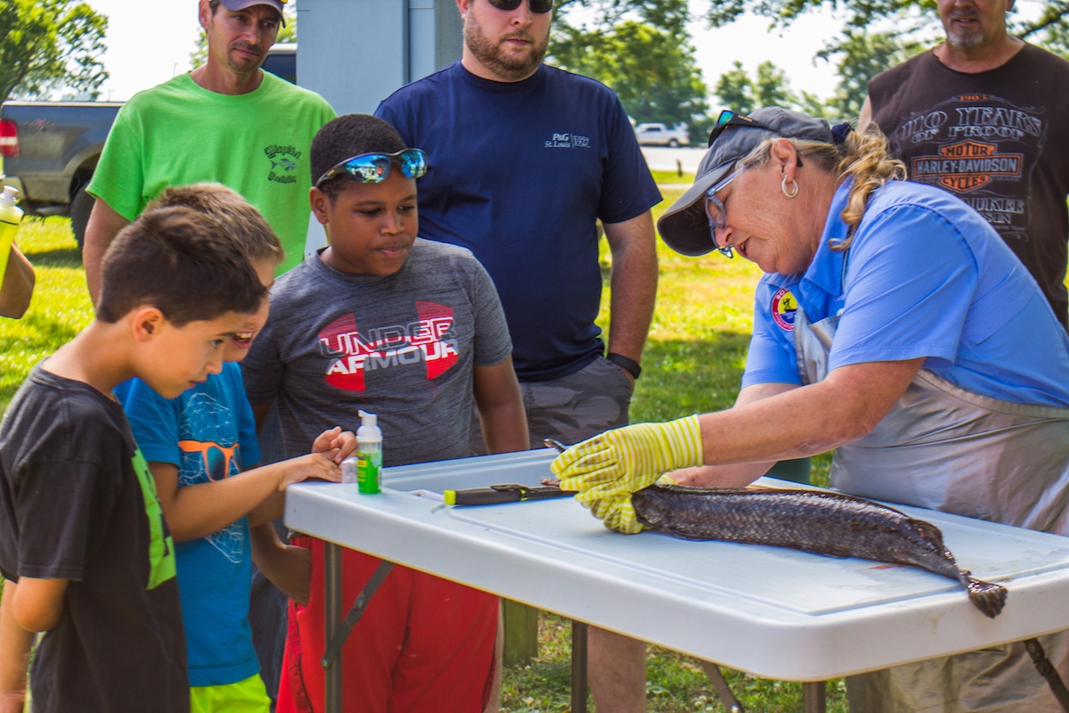 Three young boys and men in the background watch as a woman teaches all how to clean a fish for the table. In the background is a sun dappled lawn with trees and a parking lot.