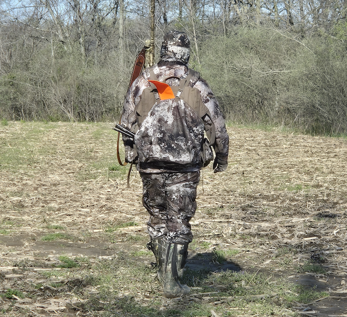 A hunter in full camouflage gear walks through an agricultural field toward a brushy edge of a woodland.