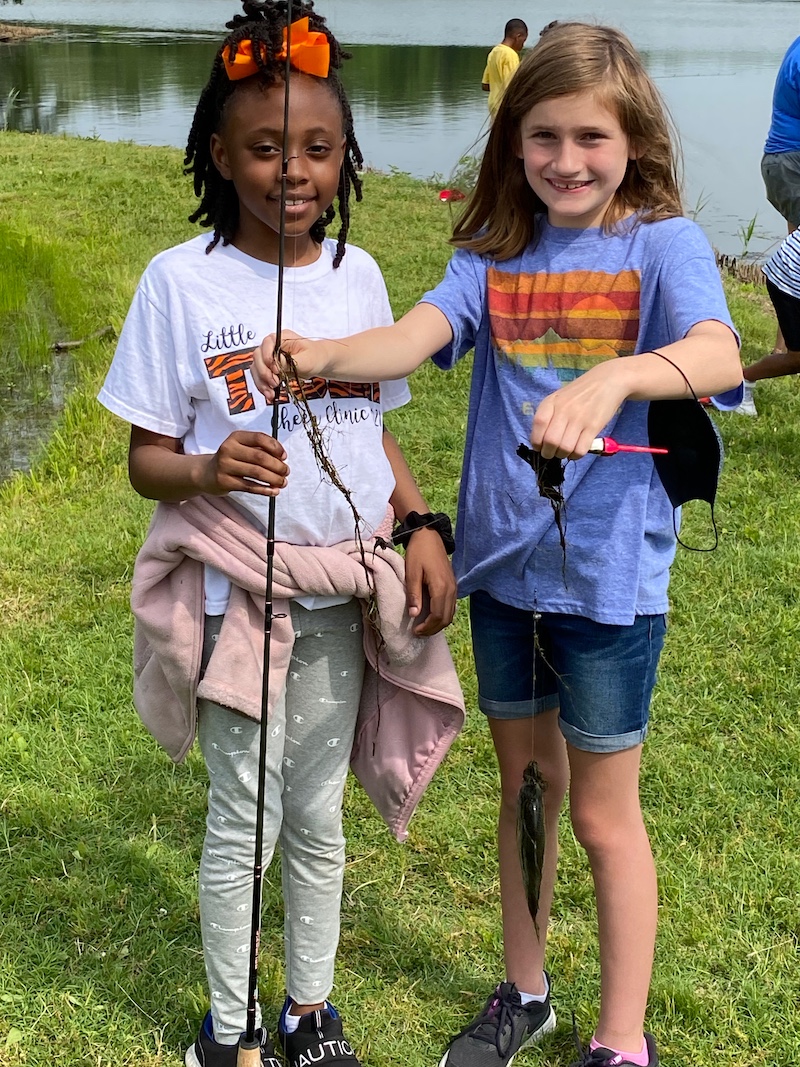 Two young girls hold a fishing rod and reel. One of the girls holds up the fishing line with a small green fish caught on the hook. In the background are other people fishing on a freshwater lake.