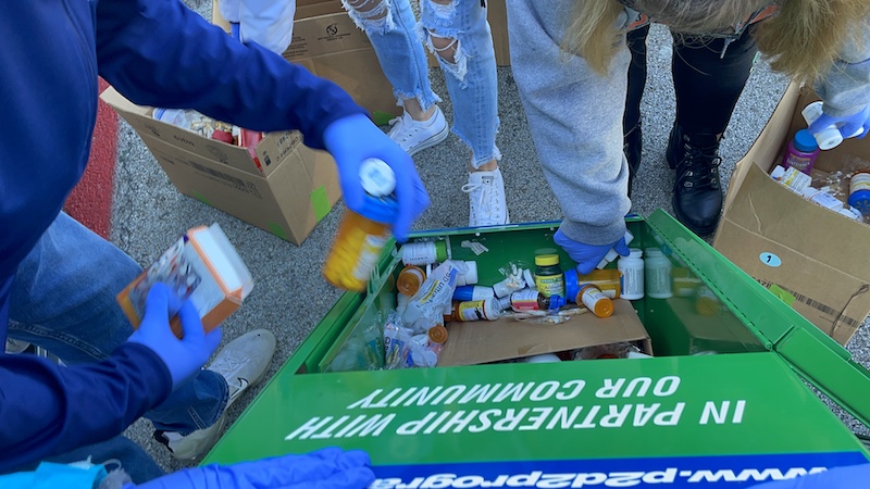 A close up of peoples arms as they remove different bottles and boxes of prescription pills and over-the-counter medicines from a green public drop-off cabinet.