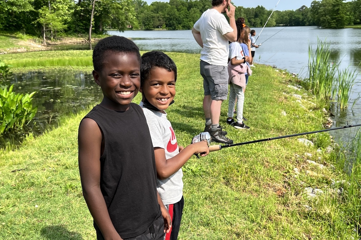 Two young boys stand together on the bank of a freshwater lake. One of the boys holds up a fishing rod and reel. In the background are a group of children and adults fishing.