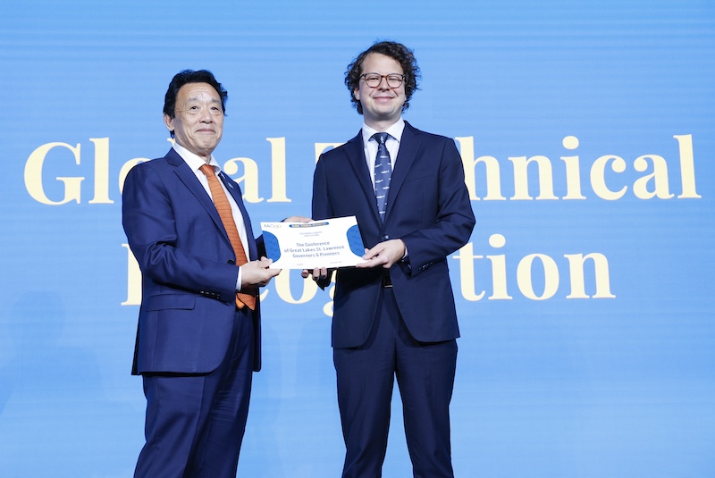 Two gentlemen wearing suits stand together and hold up a paper award certificate. In the background is a bright blue screen.