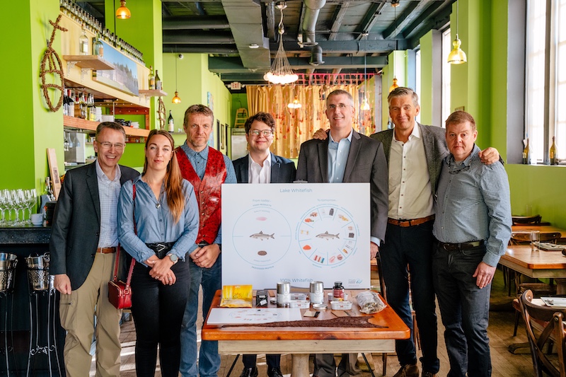 A group of people stand together posing for a photo in a restaurant with chartreuse green walls. The people stand behind a table with different fish products and a poster explaining the different products.