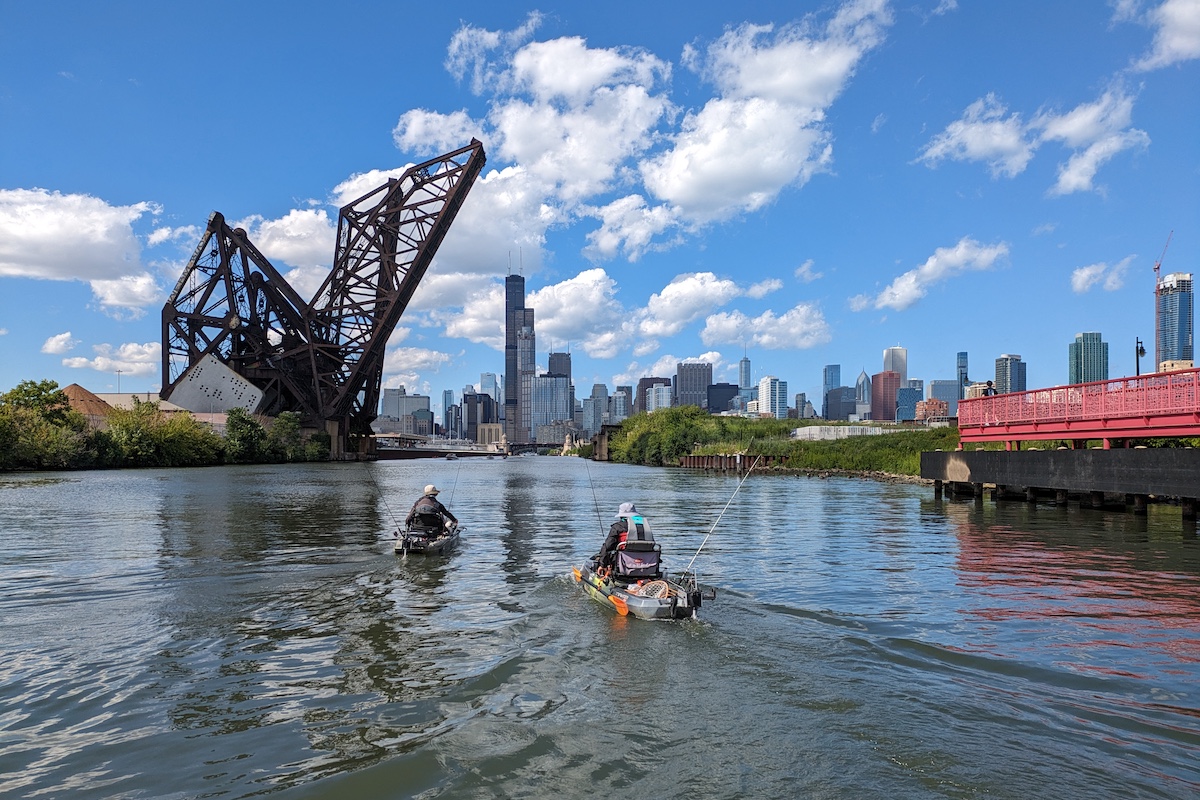 Two people floating in kayaks on a river. Each kayak has two fishing rods and reels secured upright to the sides of the boats. In the background is the Chicago skyline. A bright blue partly cloudy sky is overhead.