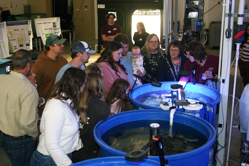 A group of people stand around three blue tanks of water. One person stands over one tank and talks to the crowd while pointing out something in the tank. Two people are in the background near some scientific research posters.