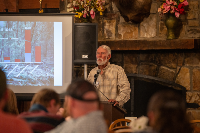 A elderly man stands at a podium with a microphone attached and speaks to a group of people. In the background is a large fireplace and projector screen with a bar graph on it.