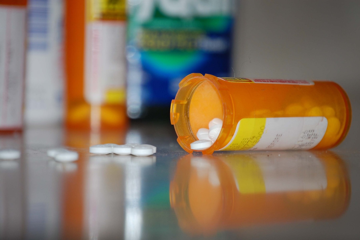 A close-up on a counter-top of an orange prescription bottle on its side with a few white capsules fallen out onto the counter outside of the bottle. In the background are more medicines and pill bottles.