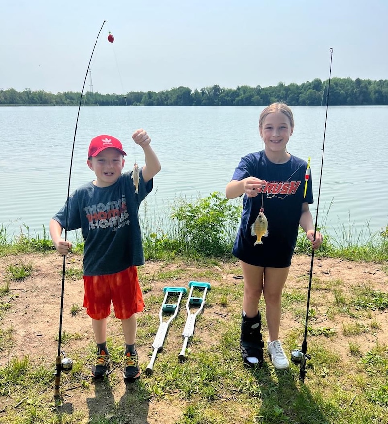 A boy and girl both holds up a fishing rod and reel in one hand and holds up the fishing line each with a small green fish caught on the hook. In the background is a freshwater lake with trees filling the bank.