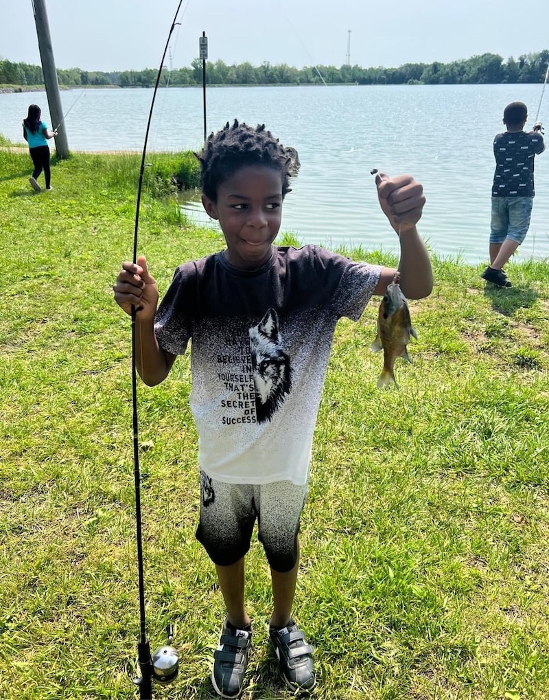 A young boy holds a fishing rod and reel in one hand and in the other he holds up a fishing line with a small green fish caught on the hook. In the background are two other children fishing on a freshwater lake.