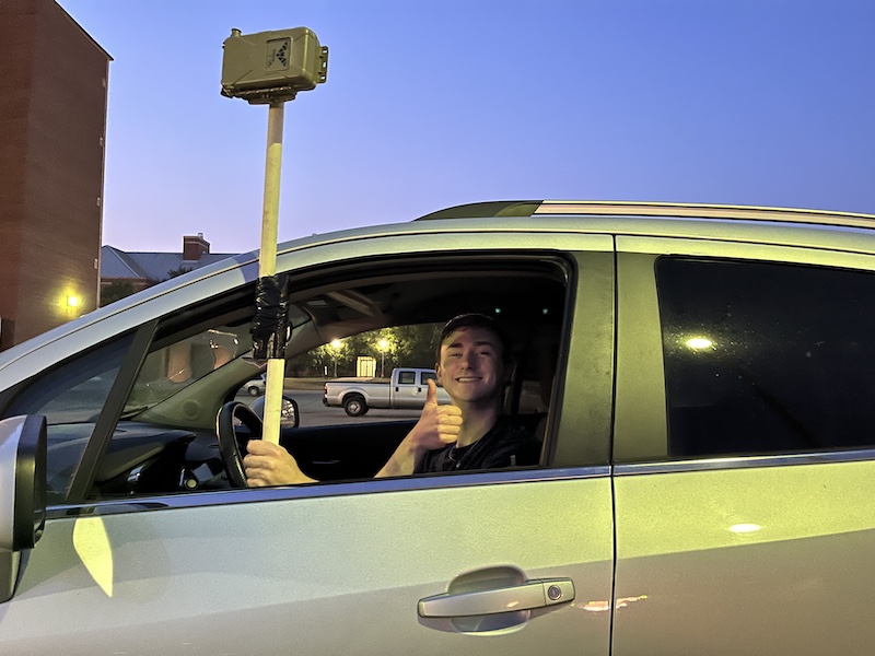 A researcher holds a brown rectangular recording device attached to a pole while sitting in the driver's seat of a car. In the background is a parking lot. 