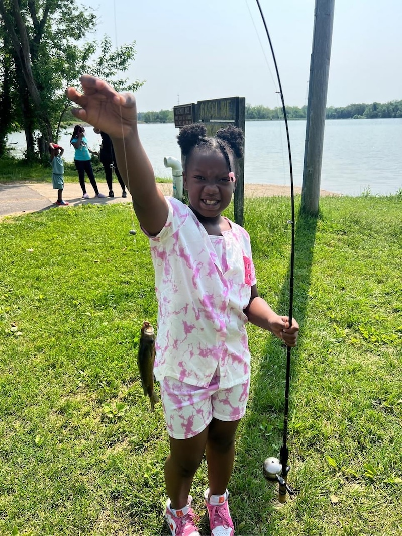 A girl holds up a fishing rod and reel with one hand and in the other hands holds the fishing line up with a small green fish caught on the hook. In the background is a freshwater lake.