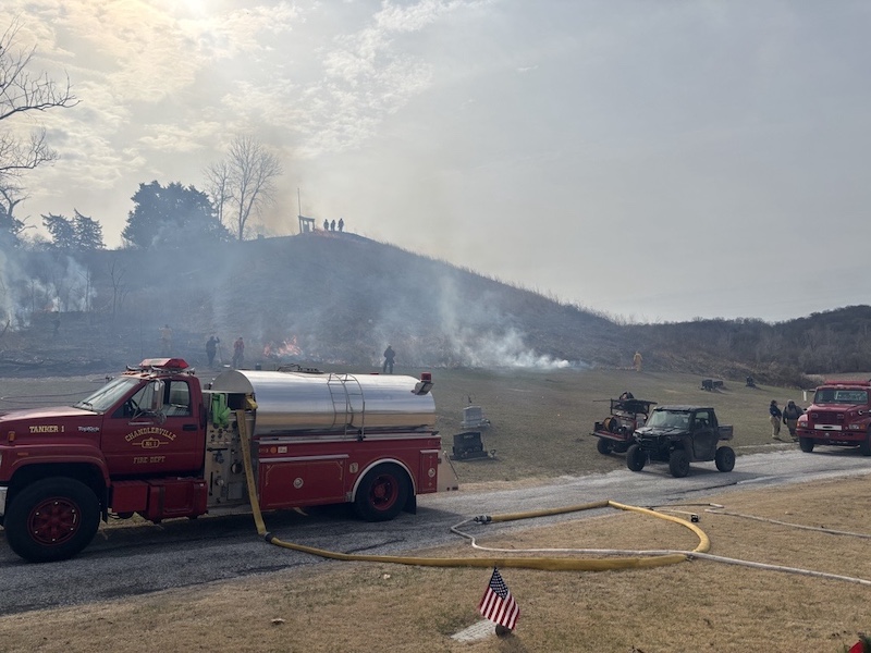 A red fire truck with an extended yellow hose, multiple all-terrain vehicles, and another large red truck park along a rural road near a hill prairie where a prescribed burn is being conducted. Several people stand around a smokey hazy prairie. Small orange flames can be seen in two places in the prairie.