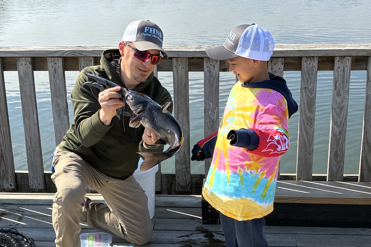 A man holds up a catfish to show a young boy. Both are standing on a wooden pier overlooking a pond.