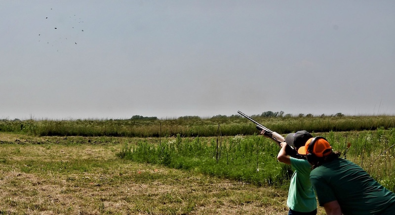 One individual aims a shotgun at a clay pigeon and pulls the trigger. Another individual stands nearby and coaches the person through the process. In the background is a partially mowed grassland against a bright blue sky.