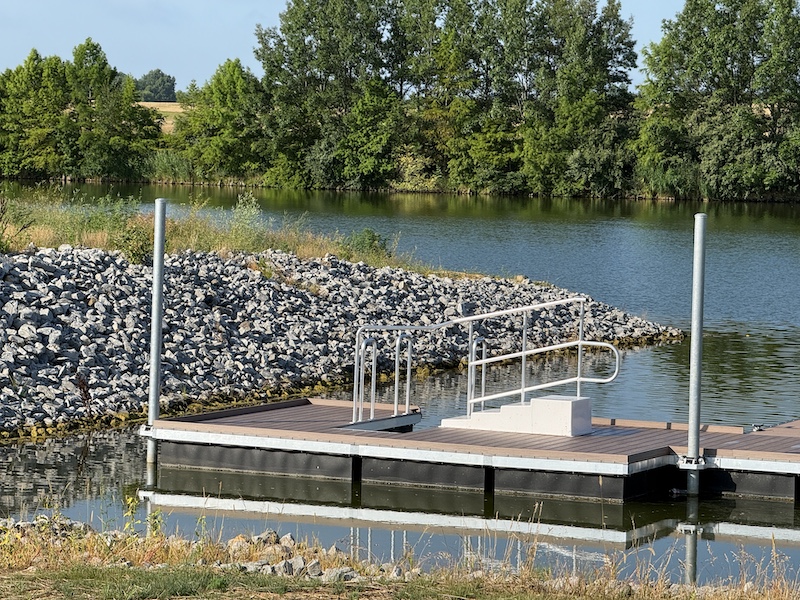 A dock on a freshwater lake. In the background is a rocky shoreline. A trees fill the shoreline in the distance.