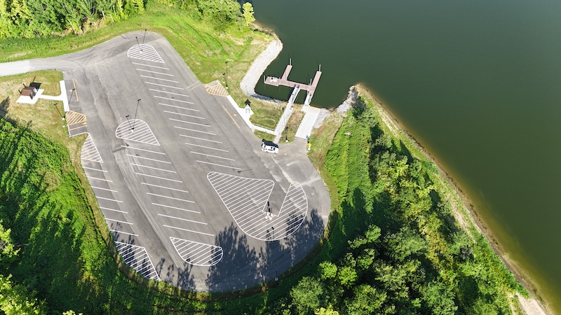 An aerial view of a parking lot, boat ramp and dock on a freshwater lake. The parking lot is surrounded by green woodlands.