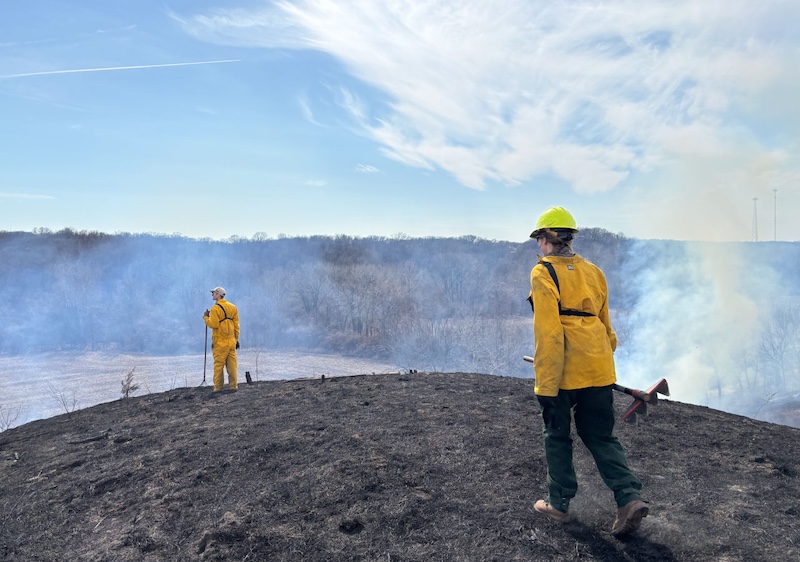 Two people wearing flame resistant gear stand on a black gray ash covered hill-top where a prescribed burn was just conducted. Smoke rises from the valley below. In the background is an agricultural field surrounded by woodlands against a bright blue partly cloudy sky.